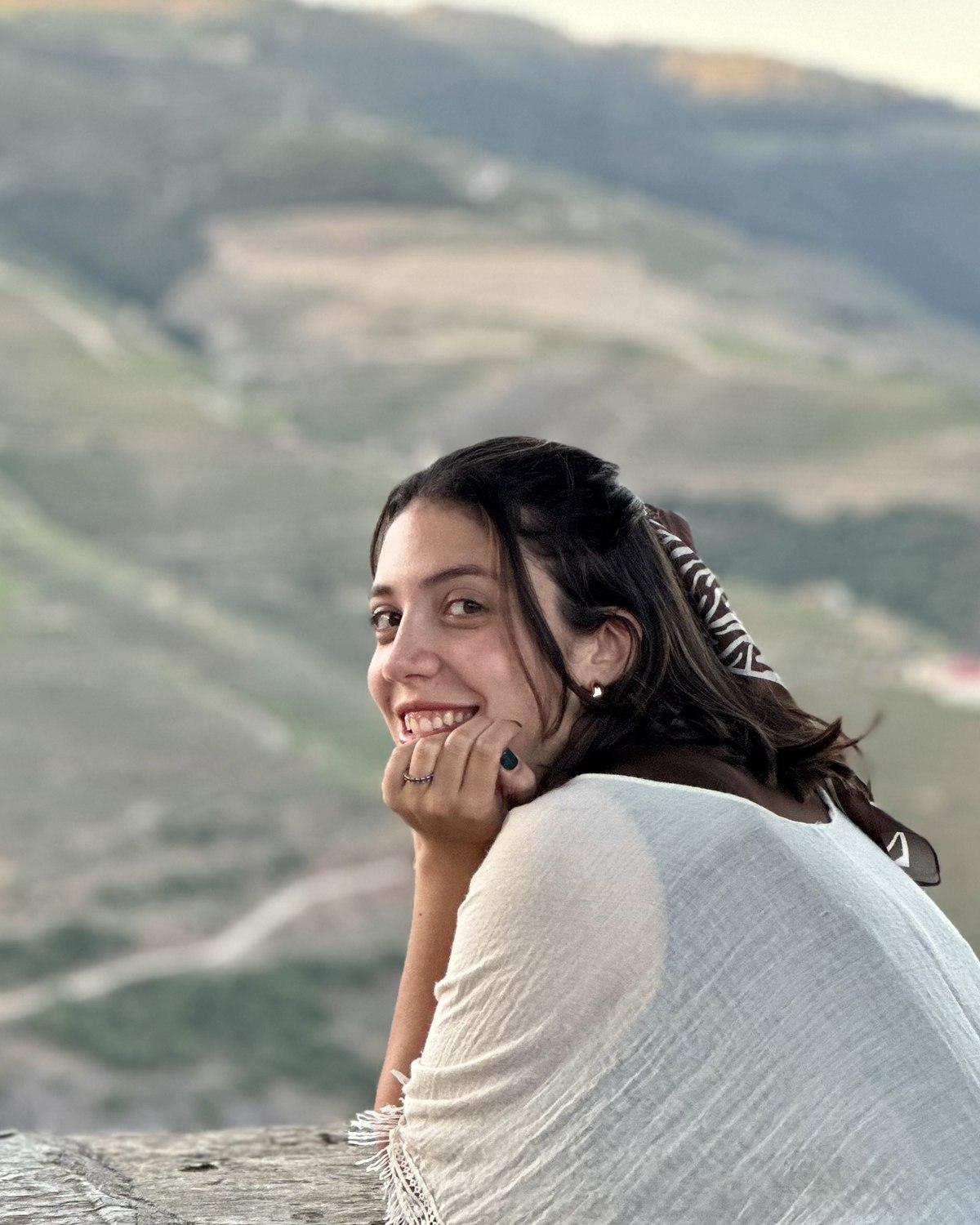 Giovanna Real sorrindo, apoiada em um muro de pedra, com montanhas ao fundo